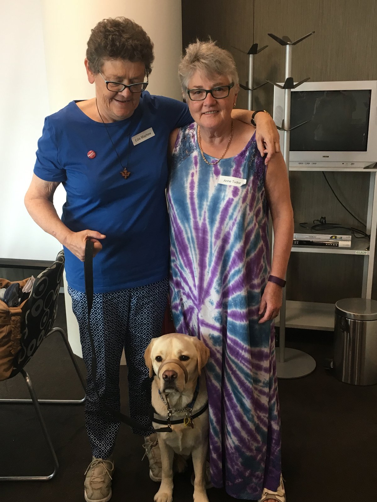 Edie Mayhew and Anne Tudor standing together with their dementia assistance dog. Edie wears a blue shirt and Anne a purple tie-dye dress.