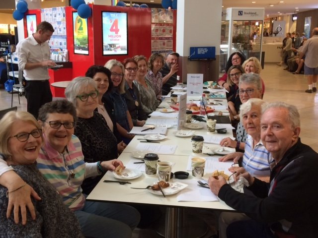 A long table at a Bigger Hearts Carers Catch Up gathering. Around a dozen people are seated together eating cake and coffee, smiling at the camera.